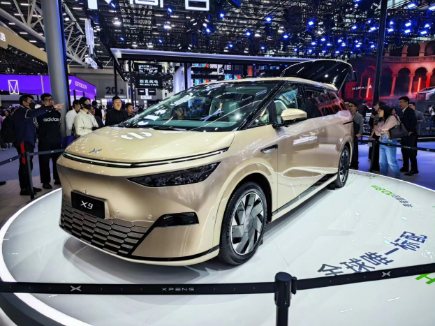 Das Xpeng X9 Auto auf einer Automesse, umgeben von Menschen, mit Pfosten, einem Zaun, einem Display, einem Gebäude und einem Dach mit Deckenleuchten im Hintergrund.