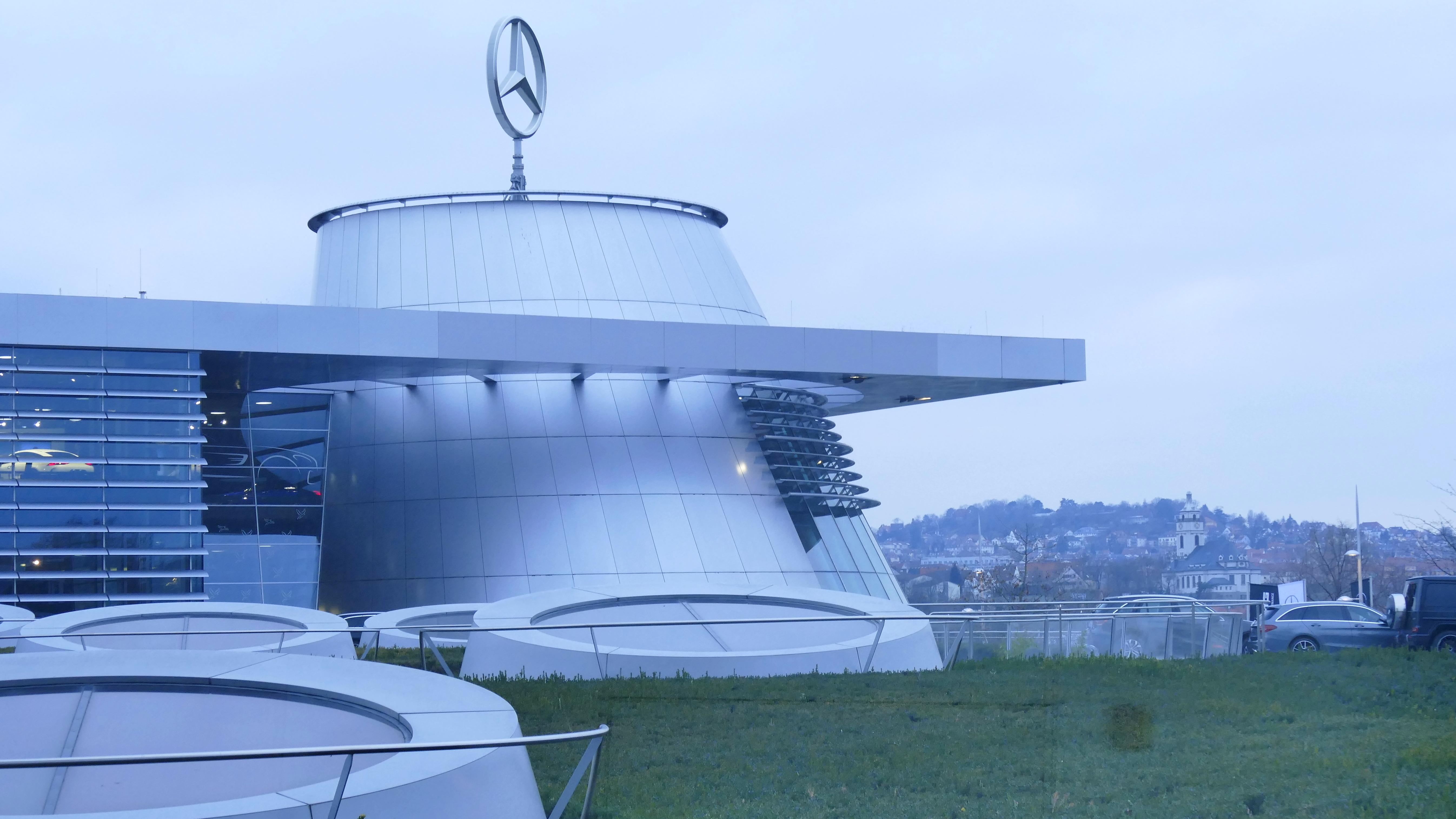 Das Mercedes-Benz Museum in Frankfurt, Deutschland - ein großes Gebäude mit Logo darauf, umgeben von Gras, Geländern, Fahrzeugen auf der Straße, Bäumen und einem klaren blauen Himmel.