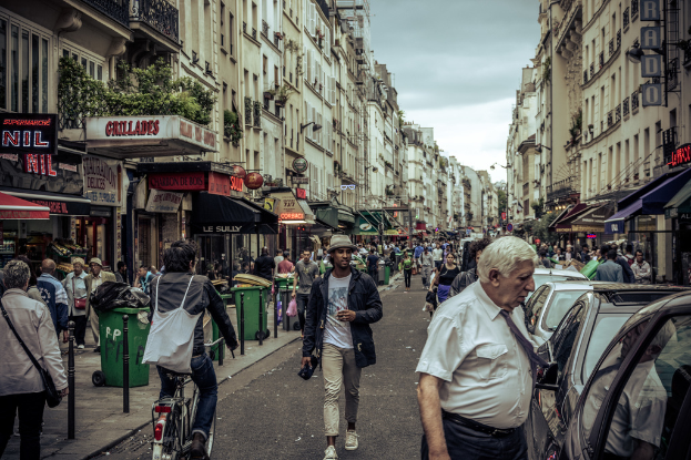 Zwei große Gebäude mit einer Straße dazwischen, wo eine Menge Leute geht und mehrere Stände auf dem Erdgeschoss eingerichtet sind, mit einigen Mülltonnen sichtbar. Ein Mann fährt ein Fahrrad auf der Straße, und Autos sind auf der gegenüberliegenden Seite geparkt.