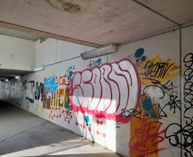 Eine mit Graffiti bedeckte Wand in einer Tiefgarage, beleuchtet von Deckenlampen, die eine Vielfalt an farbenfroher Straßenkunst zeigt.