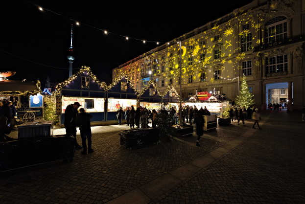 Ein geschäftiger Weihnachtsmarkt in Berlin, Deutschland mit Menschen, die sich um geschmückte Stände und festlich geschmückte Gebäude unter einem dunklen Himmel versammeln.