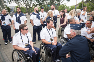 Eine Gruppe von Menschen im Rollstuhl unterhält sich vor einer Kulisse aus Bäumen, Pfählen, Gebäuden und einem klaren blauen Himmel, wobei einige Mützen und Brillen tragen, was darauf hindeutet, dass sie an den Paralympischen Spielen teilnehmen.