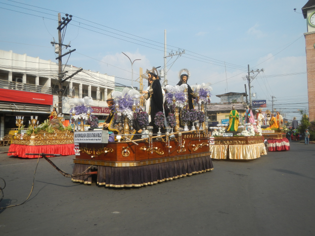 Ein Paradewagen mit Menschen darauf, umgeben von Gebäuden, Strommasten mit Drähten, Laternenmasten, Bäumen und einem Kirchturm im Hintergrund unter einem bewölkten Himmel.