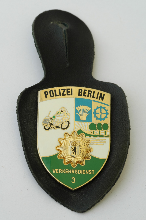 Ein schildförmiges Polizeibadgebnis mit "Polizei Berlin" in schwarzer Schrift auf einem blauen Hintergrund, das ein weißes Stern- und Rahmen-Design hat und auf einer Oberfläche liegt.