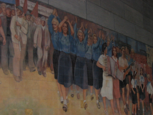 Eine Gruppe von Menschen mit blauen-kleideten Mädchen in der Mitte, Musikern auf der rechten Seite, spielenden Kindern in der rechten Ecke und Individuen mit roten Fahnen auf der linken Seite.