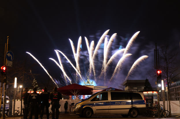 Polizeifahrzeug vor einer Menschenmenge mit farbigen Feuerwerken am Himmel und beleuchteten Gebäuden und Bäumen im Hintergrund.