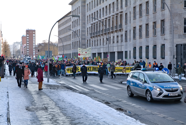 Eine große Gruppe von Menschen marschiert auf einer verschneiten Straße in Berlin, Deutschland, mit Schildern und Plakaten, mit Gebäuden, Bäumen und einem geparkten Auto im Hintergrund.