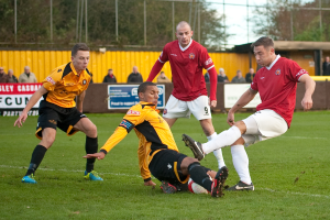 Zwei Spieler in weißen Uniformen spielen auf einem Rasenfeld mit einem Ball, während Spieler in roter Uniform im Hintergrund sind. Viele Zuschauer stehen außerhalb des Feldes und feuern die Spieler an, mit einem Baum und dem Himmel im Hintergrund.