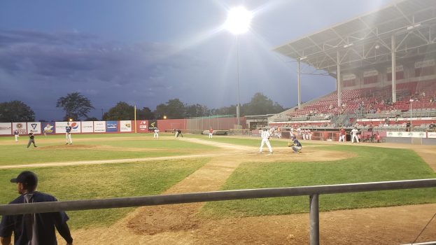 Baseballspiel in einem Stadion mit Zuschauern auf den Rängen, Bäumen und Stadioninfrastruktur im Hintergrund unter einem klaren Himmel.