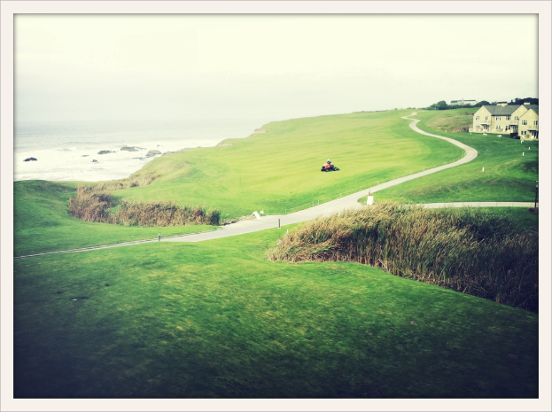 Landschaftsszene mit Unkraut und Feldern im Vordergrund, einem Traktor und einem Weg in der Mitte, Häusern und Bäumen oben rechts, Wasser oben links und einem bewölkten Himmel.