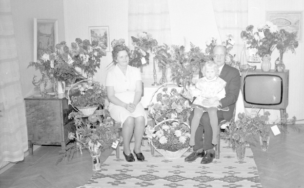 Schwarzes und weißes Foto einer Familie aus drei Personen - ein Mann hält ein Kind, eine Frau und das Kind sitzen auf einem Sofa in einem Wohnzimmer mit Blumenvasen, einem Teppich, gerahmten Wanddekorationen und Vorhängen.