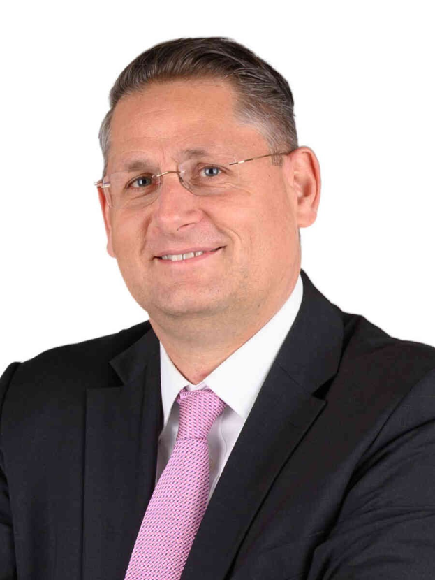 Ein lachender Mann im Anzug und mit Krawatte, der eine Brille trägt, vor einem weißen Hintergrund.