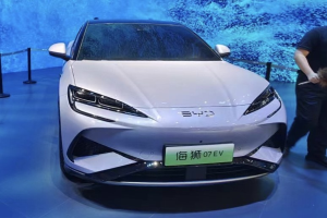 Ein weißes Auto auf einer Automesse mit einer Person rechts daneben, die ein Objekt hält, ein Bildschirm im Hintergrund und Absperrpoller mit Seilen links.