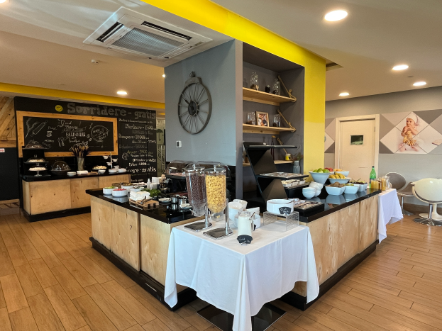 Ein Restaurantinnenraum mit Tischen und Stühlen, Essensbehältern, einem Schwarzen Brett mit Text, einer Wanduhr, Deckenleuchten, einer Klimaanlage und einer Tür im Hintergrund, das sich im Hôtel de Ville in Paris befindet.