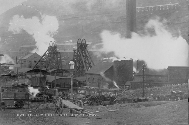 Schwarzes und weißes Foto einer Industrieanlage, die Rauch ausstößt, umgeben von Gebäuden, Strommasten, Fahrzeugen, Karren und Text am unteren Rand.