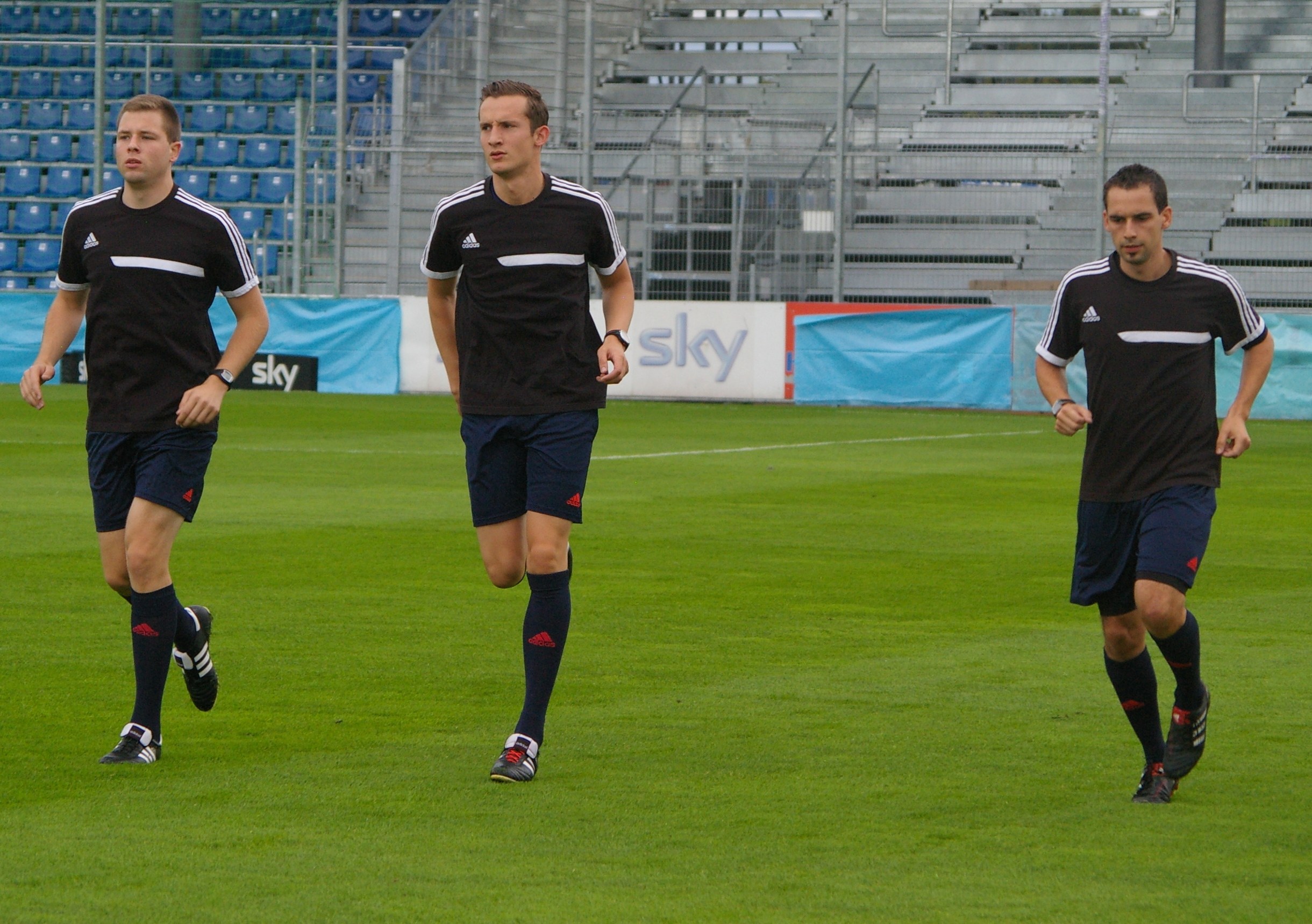 Drei Männer in schwarzer Sportbekleidung auf einem Fußballfeld mit Tribünen und Bannern im Hintergrund.