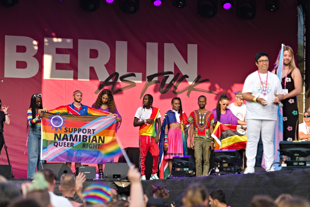 Eine Gruppe von Menschen auf einer Bühne mit einem Banner, auf dem 'Berlin als Fuck' steht, mit Lautsprechern, Mikrofonen und einer Menge darunter, die ihre Hände hebt.