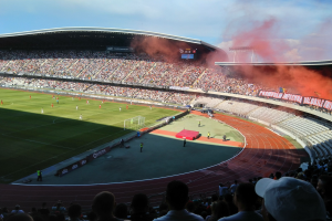 Ein großer Stadion mit Zuschauern, die ein Fuballspiel verfolgen, mit Spielern im Einsatz auf dem Feld und Text am unteren Rand des Bildes.