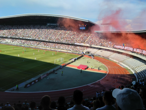 Ein großer Stadion mit Zuschauern, die ein Fuballspiel verfolgen, mit Spielern im Einsatz auf dem Feld und Text am unteren Rand des Bildes.
