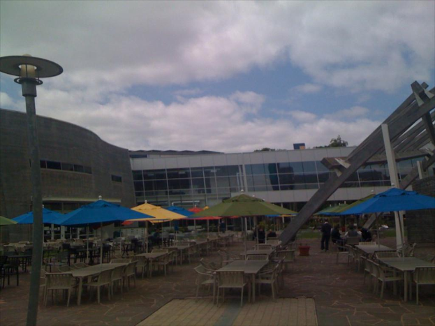 Ein Resort-Szenario mit Stühlen, Tischen, Sonnenschirmen und einer Lampe im Vordergrund, Bäumen und Gebäuden in der Mitte, unter einem bewölkten Himmel.