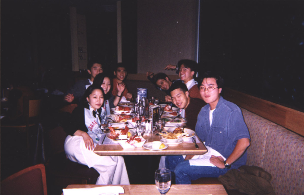 Eine Gruppe von Menschen sitzt um einen Tisch, der mit verschiedenen Speisen, Tellern, Karten, Gläsern, Taschentüchern und Besteck gefüllt ist, mit einer Jacke auf der rechten Seite, einem weiteren Tisch mit einem Glas davor, einem Stuhl auf der linken Seite und einem Fenster in der Mitte.