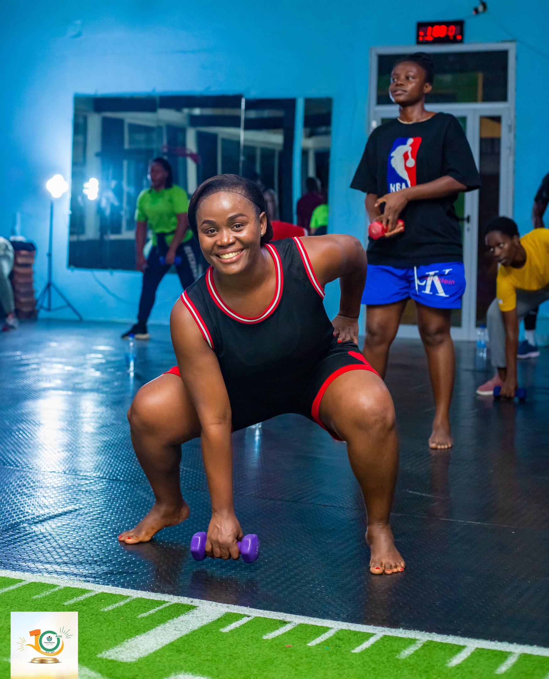 Eine Gruppe von Menschen in einem Fitnessstudio mit einer Frau, die Kniebeugen macht, während sie Hanteln hält, umgeben von Equipment und Branding für ein Fitnessprogramm.