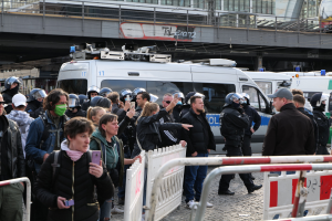 Eine Gruppe von Menschen vor Polizeifahrzeugen mit einigen, die Helme tragen und Handys halten, hinter Absperrungen, mit einer Brücke und Gebäuden im Hintergrund während einer Demonstration in Berlin.