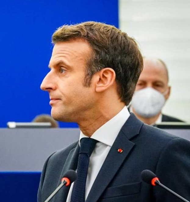 Ein Mann im Anzug und Krawatte steht an einem Podium mit zwei Mikrofonen, während eine andere Person mit einer Maske dahinter steht, mit einem unscharfen Hintergrund.