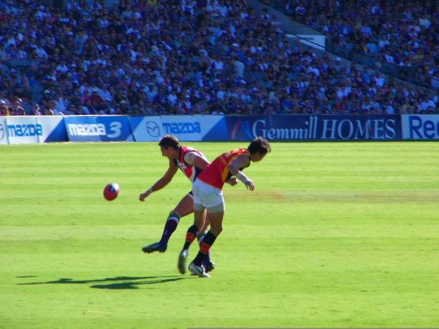 Zwei Personen in rot-weißer Kleidung stehen mit einem Ball in der Luft, während Banner und eine Zuschauermenge im Hintergrund des Stadions zu sehen sind.