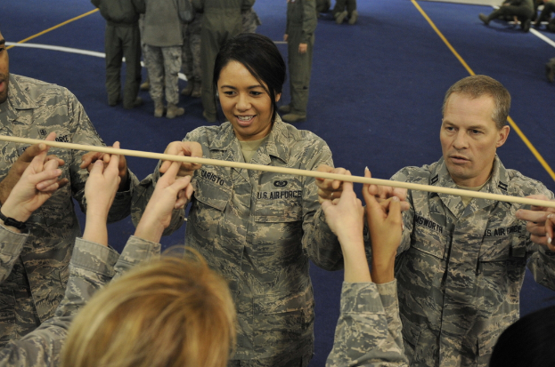 Mehrere Personen in Armeeuniformen stehen, wobei eine einen Stock hält, während andere sitzen oder stehen und im Hintergrund ein blauer Boden zu sehen ist.