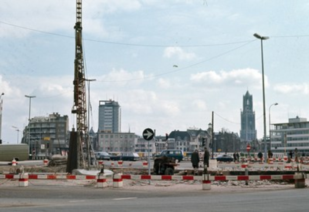 Bauplatz mit einem zentralen Turm, umgeben von Gebäuden, Straßeninfrastruktur, Fahrzeugen und bewölktem Himmel.