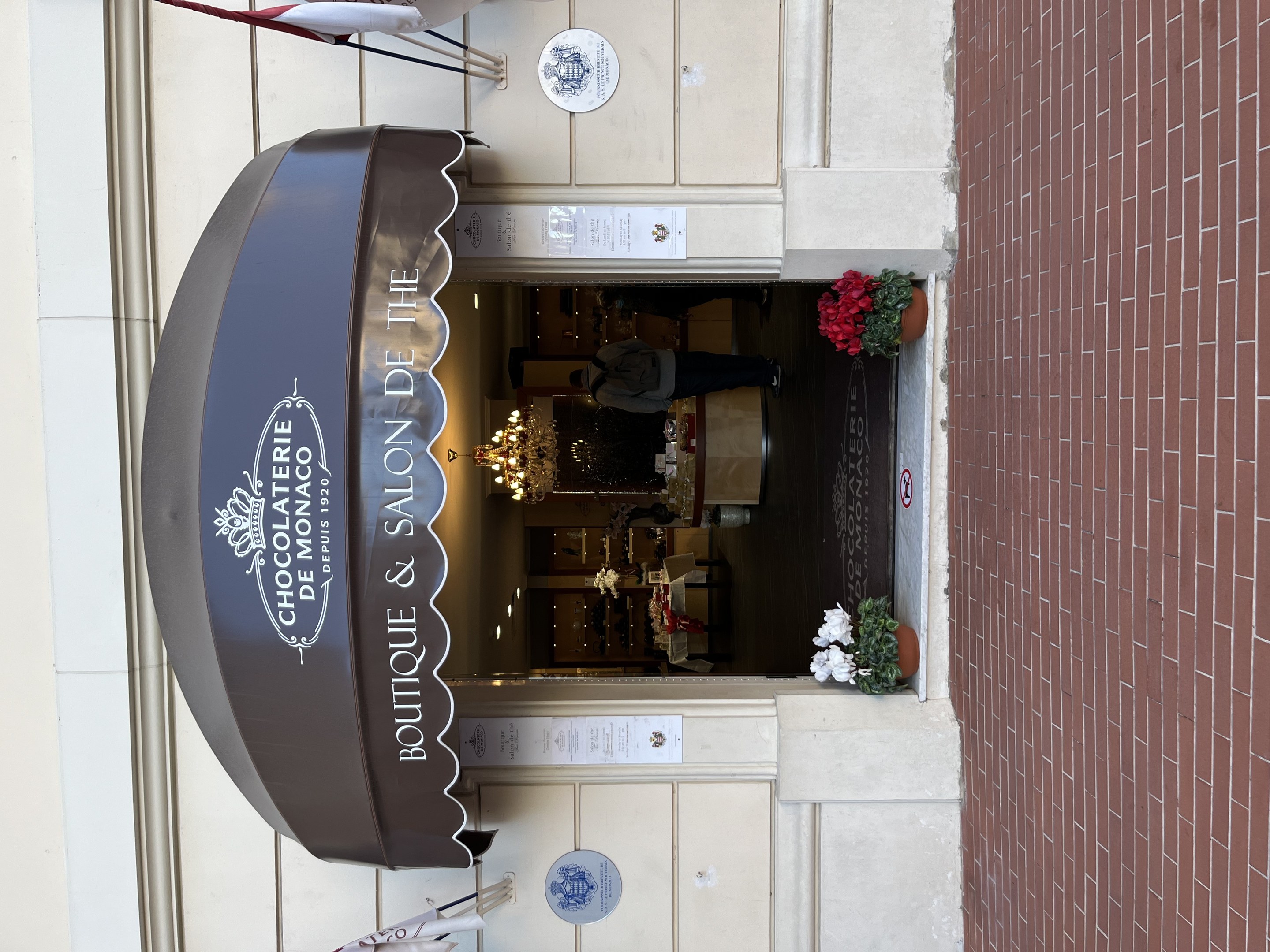 Außenansicht eines Gebäudes mit einer "Boutique & Salon de Monaco"-Tafel, das Sitzgelegenheiten, Blumentöpfe und eine sichtbare Inneneinrichtung mit Beleuchtung und Dekoration zeigt.