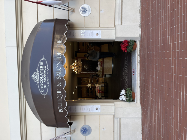 Außenansicht eines Gebäudes mit einer "Boutique & Salon de Monaco"-Tafel, das Sitzgelegenheiten, Blumentöpfe und eine sichtbare Inneneinrichtung mit Beleuchtung und Dekoration zeigt.