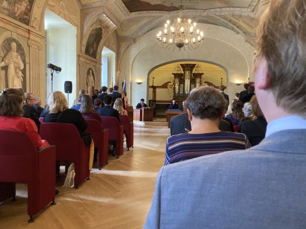 Eine Gruppe von Menschen sitzt in Stühlen in einem Raum mit Rahmenfotos an der Wand und einer Kristallleuchter, die an einer Besprechung im schwedischen Parlament mit einem Podium, Mikrofon und Flaggen im Hintergrund teilnehmen.
