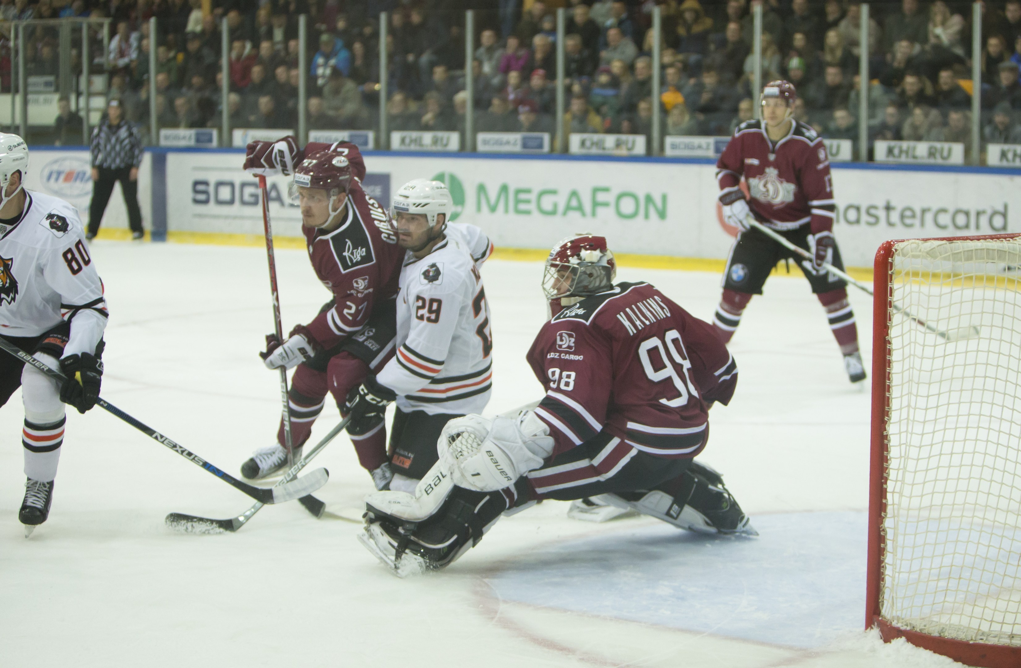 Gruppe von Menschen, die Hockey auf einem Eisring spielen, mit Tor auf der rechten Seite, Helm und Stöcke tragend, Zuschauer in Tribünen mit Bannern im Hintergrund.