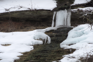 Ein kleiner Wasserfall ergießt sich über einen schneebedeckten, vereisten Felsen in einem bewaldeten Gebiet, umgeben von schneebedeckten Bäumen.