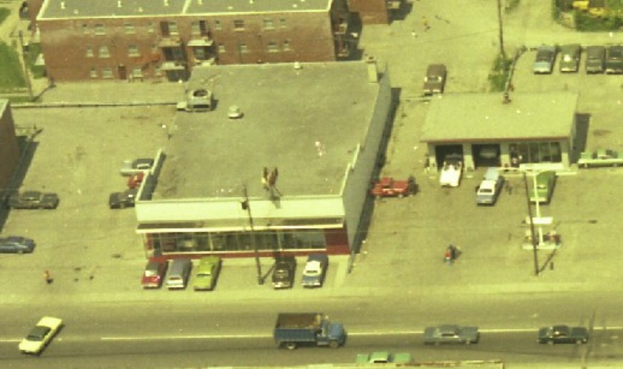 Luftaufnahme einer Stadt mit zahlreichen parkenden Autos um Gebäude herum und einigen Autos auf der Straße.