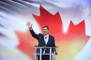 Der kanadische Premierminister Alex Salmond steht an einem Rednerpult mit Mikrofonen, lächelnd vor einer kanadischen Flagge, mit einem Bildschirm im Hintergrund.