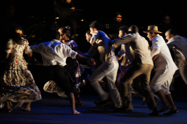 Eine Gruppe von Menschen tanzt im Vordergrund mit einer anderen Gruppe, die steht und Mikrofone im Hintergrund, alles auf einem Boden.
