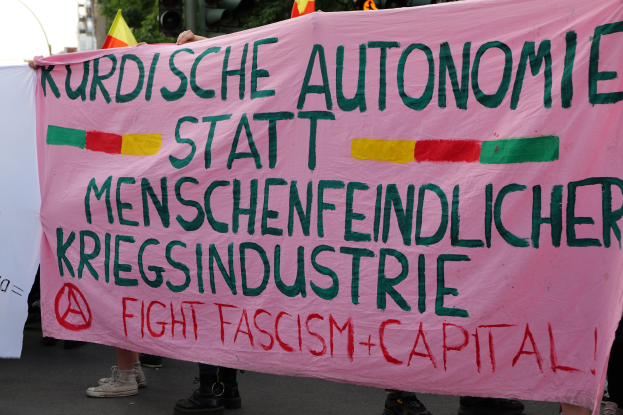 Eine Gruppe von Menschen mit einer pinken Fahne, auf der steht "Kurdische Autonomie Statt Menschenfeindlicher Kriegsindustrie Fight Fascism Capital" auf einer Straße mit Ampeln, Flaggen, Bäumen, Gebäuden und einem klaren blauen Himmel im Hintergrund.