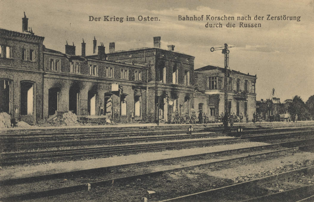 Ein altes Schwarz-Weiß-Foto eines Bahnhofs namens "Bahnhof Korschen in Ostern, Deutschland" mit Bahnschienen, Gebäuden, Pfählen, Menschen, Bäumen und einem Himmel im Hintergrund.