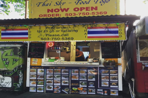 Ein Geschäft mit verschiedenen Artikeln, darunter Getränkedosen, Speisekarten, eine Uhr, Gewürze in Plastikbehältern, Kondimente und Werbetafeln unter einem klaren Himmel.