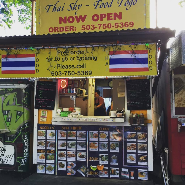 Ein Geschäft mit verschiedenen Artikeln, darunter Getränkedosen, Speisekarten, eine Uhr, Gewürze in Plastikbehältern, Kondimente und Werbetafeln unter einem klaren Himmel.