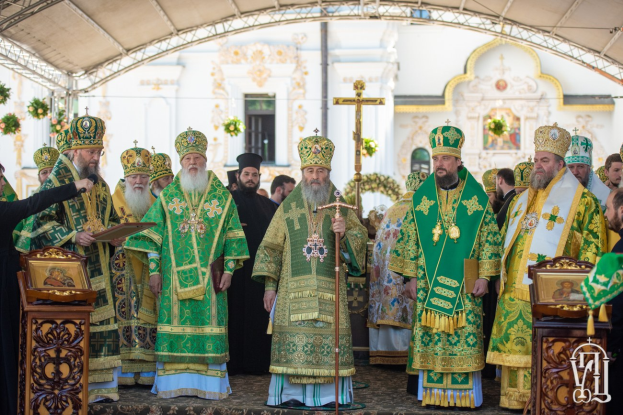 Eine Gruppe von Priestern vor einer Kirche, wobei einer ein Buch und ein Mikrofon hält, während einer religiösen Zeremonie mit einem Kreuz, Blumen und einem Gebäude im Hintergrund.