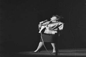 Eine Frau in einem schwarzen und weißen Outfit aus den 1950er Jahren singt fröhlich auf einer von einem Scheinwerfer beleuchteten Bühne und hält ein Mikrofon.