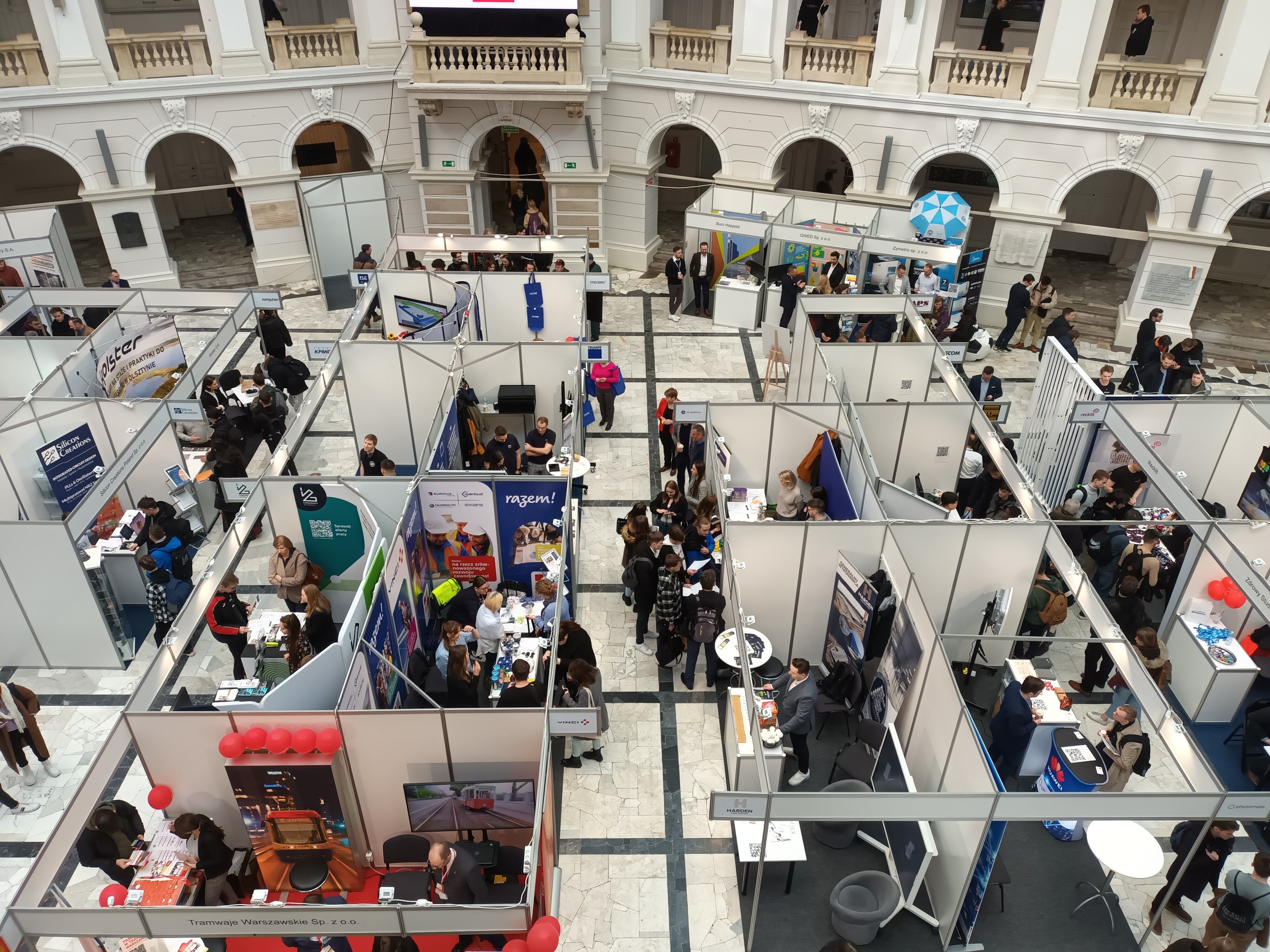Große Gruppe von Teilnehmern auf einer internationalen Messe mit Ständen, Bannern, Tischen, Stühlen und Ballons in einem Raum mit architektonischen Bögen, Säulen und einem Bildschirm im Hintergrund.