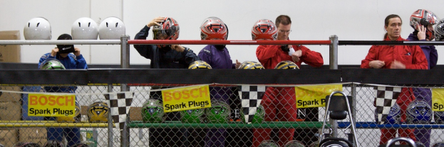 Ein Maschendrahtzaun mit Plakaten und Fahnen, mit ein paar Menschen im Hintergrund, einige tragen Helme, und zusätzliche Helme auf einem Ständer, alles vor einer Wand.
