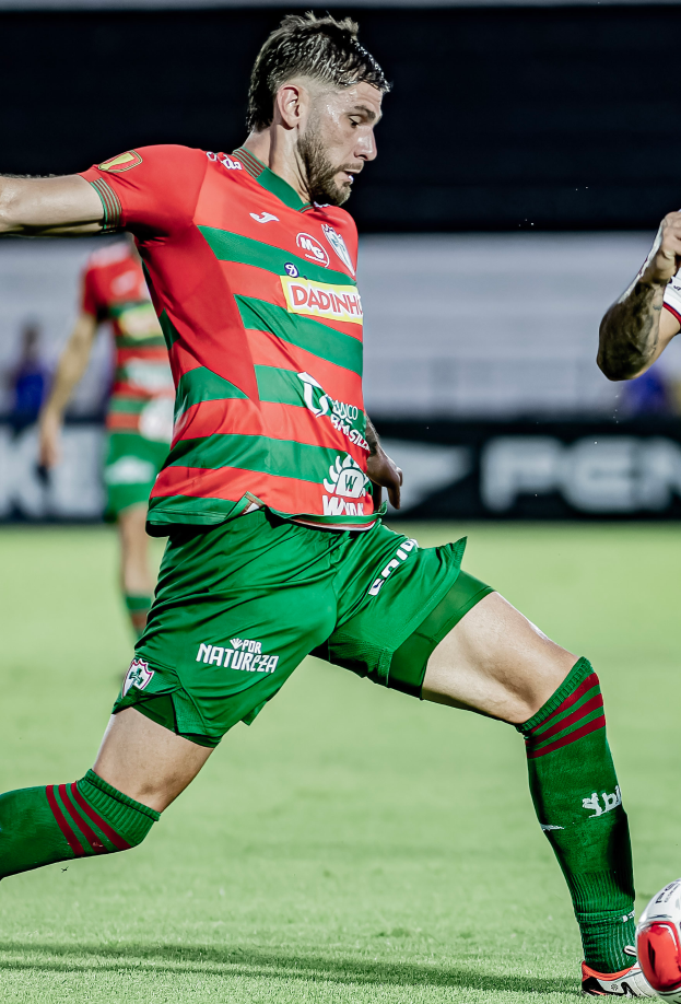 Ein Mann in roter und grüner Uniform kickt einen Fußball auf einem Rasenfeld, mit einer anderen Person im Hintergrund und einer Tafel, auf der 'Santos gegen Vitoria Guimarães' steht.