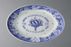 Ein blau-weißer Teller mit dem Olympischen Logo und detaillierten Designs, wahrscheinlich zur Erinnerung an die Olympischen Spiele in München, vor einem grauen Hintergrund.