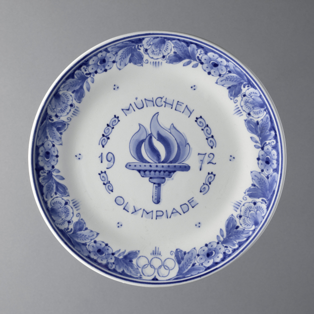 Ein blau-weißer Teller mit dem Olympischen Logo und detaillierten Designs, wahrscheinlich zur Erinnerung an die Olympischen Spiele in München, vor einem grauen Hintergrund.